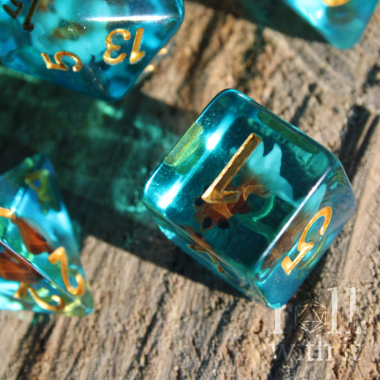 Close-up of teal dice with gold numbers and a gold fish swimming inside on a wooden surface