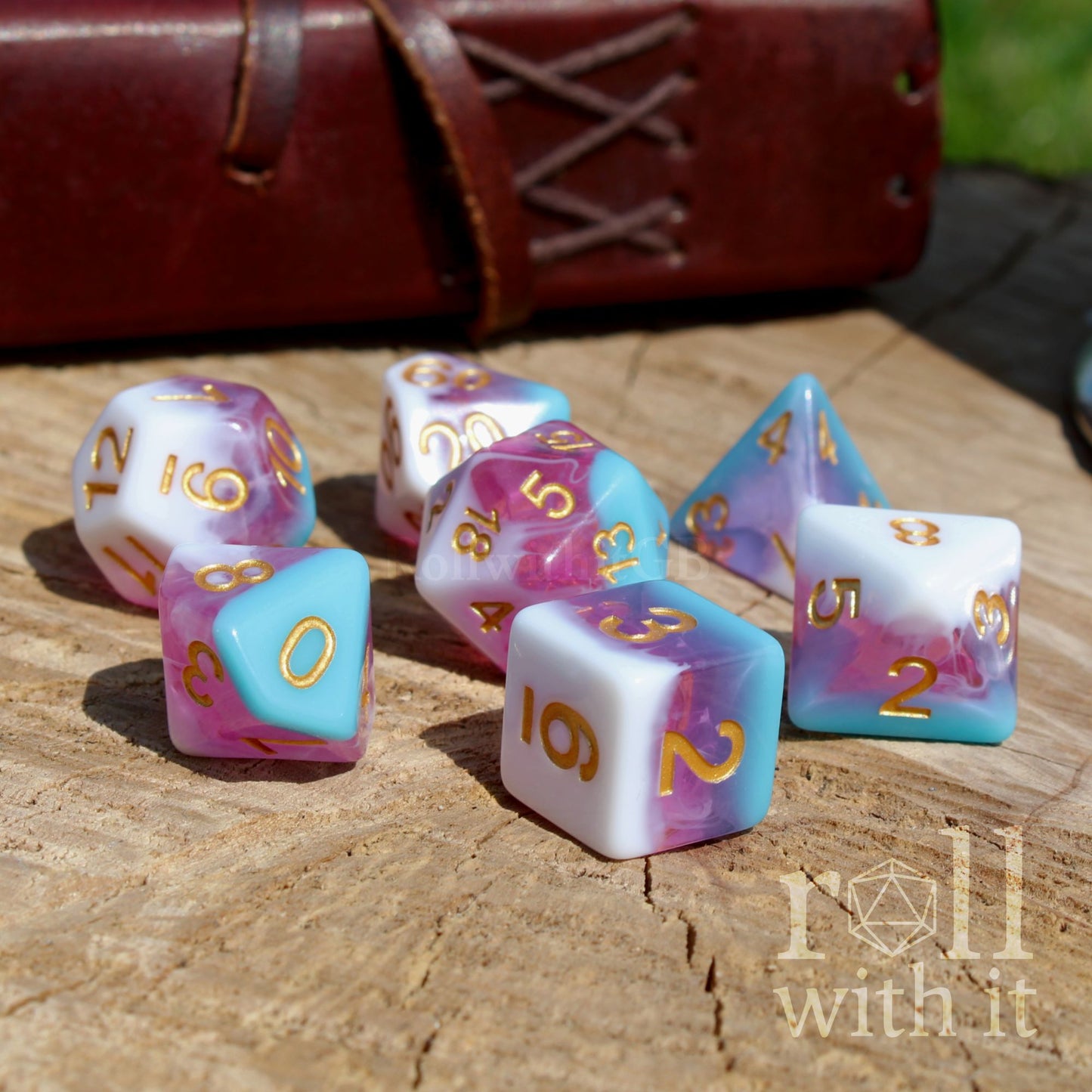 Set of colorful clear pink cloudy white blue polyhedral dice on a wooden surface with 'roll with it' branding.