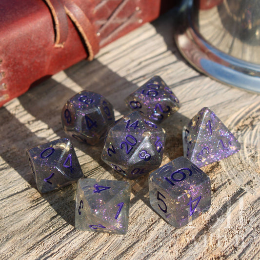 Set of purple and clear grey gold foil filled polyhedral dice on a wooden surface with a leather book in the background.