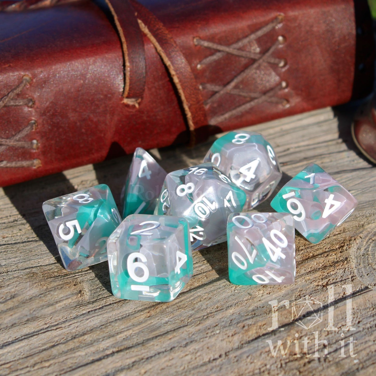 Set of teal and clear white polyhedral dice on a wooden surface with a leather-bound book in the background.