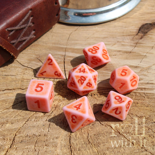 Set of bright pink polyhedral dice with red numbers on a wooden surface, featuring a leather-bound notebook.