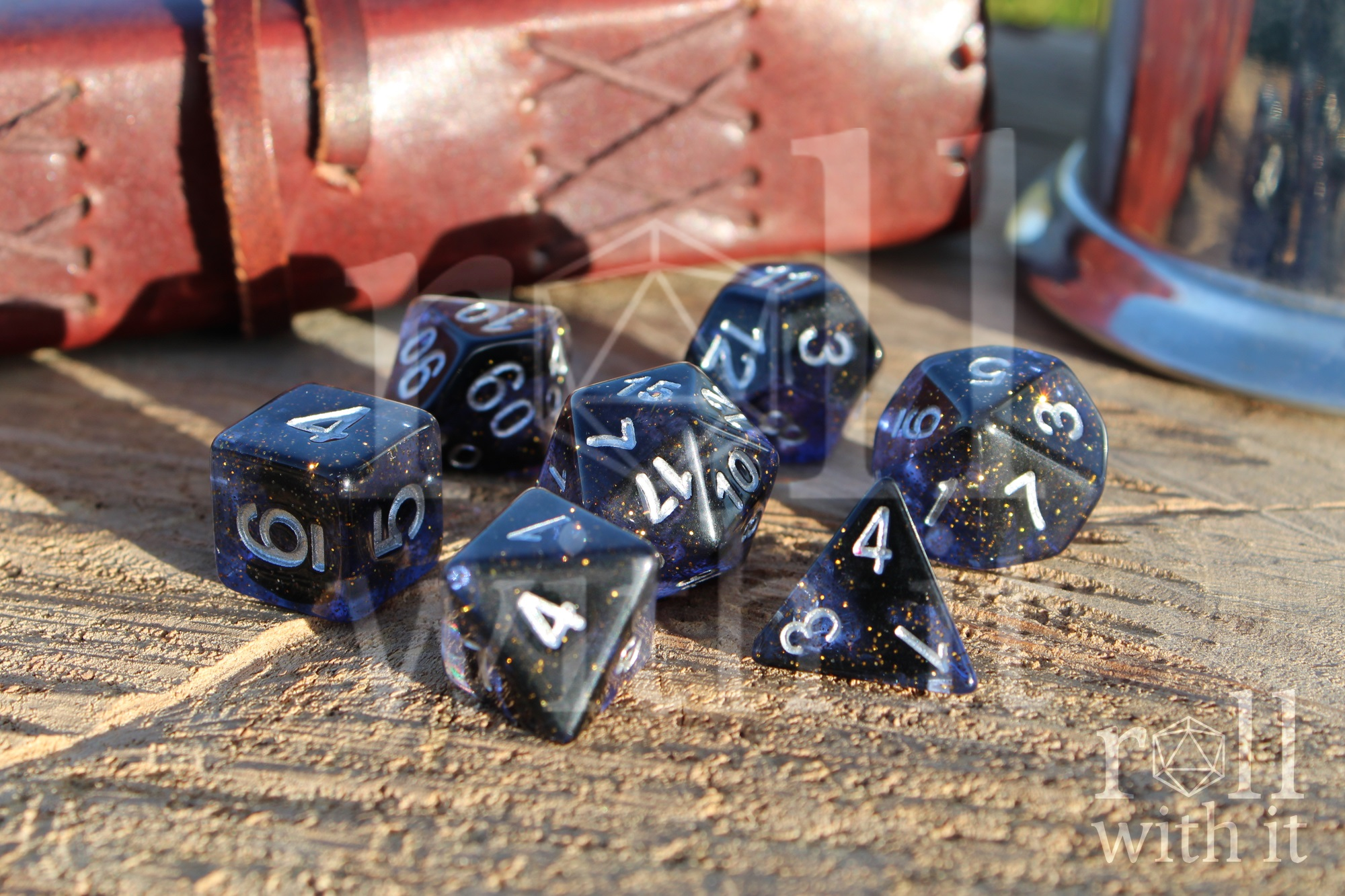 Set of deep blue polyhedral dice with gold flecks on a wooden surface with a leather journal in the background.