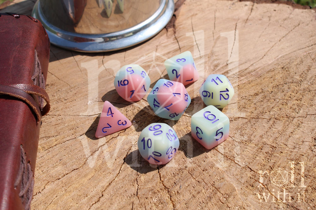 Set of colourful matt pastel polyhedral DnD dice on a wooden surface with a leather-bound journal in the background.