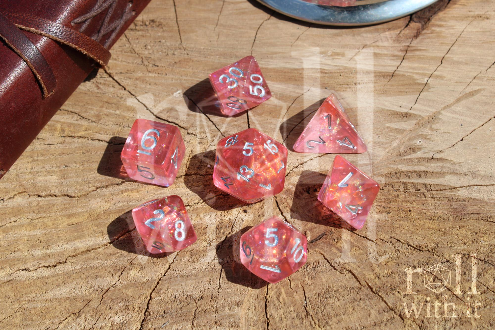 Shimmering Pink polyhedral dice set on a wooden surface with a leather-bound book.