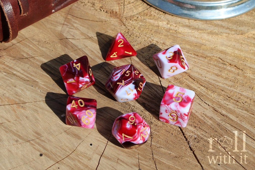 A set of 7 cherry red and swirling creamy white polyhedral TTRPG dice on a oak base with role playing game accessories in the background