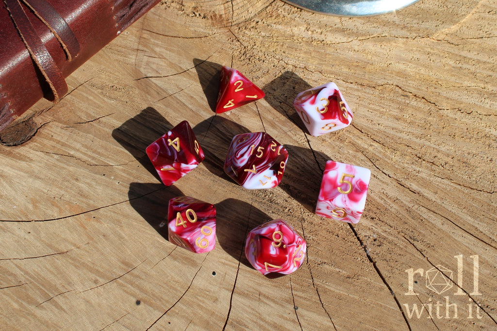A set of 7 cherry red and swirling creamy white polyhedral TTRPG dice on a oak base with role playing game accessories in the background