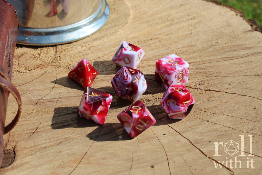 A set of 7 cherry red and swirling creamy white polyhedral TTRPG dice on a oak base with role playing game accessories in the background