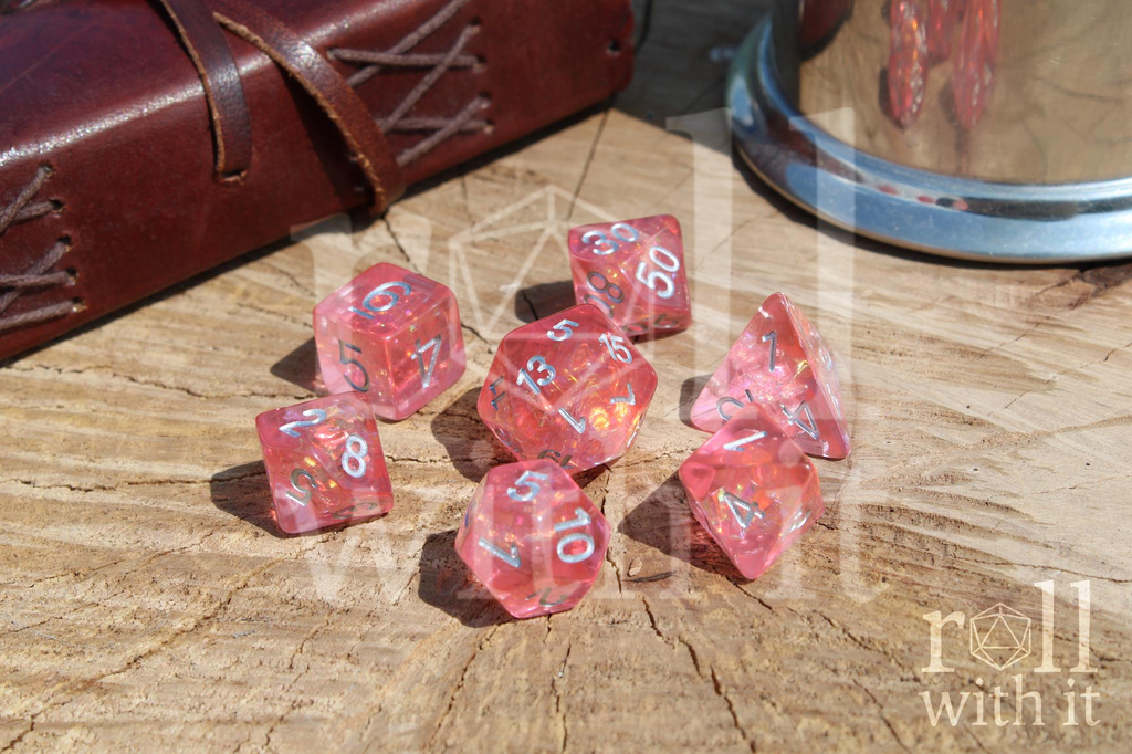 Shimmering Blossom Pink polyhedral DnD dice set with silver numbers, on a wooden surface with 'roll with it' branding.