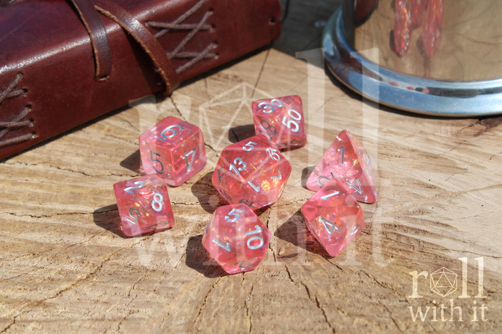Shimmering Blossom Pink polyhedral DnD dice set with silver numbers, on a wooden surface with 'roll with it' branding.