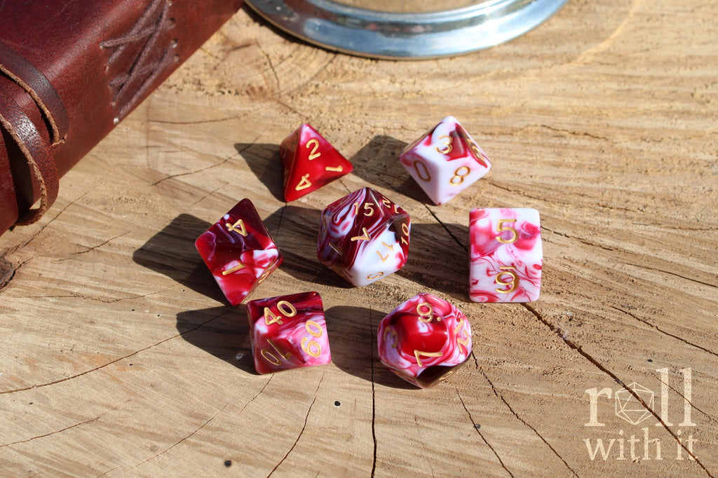 A set of 7 cherry red and swirling creamy white polyhedral TTRPG dice on a oak base with role playing game accessories in the background
