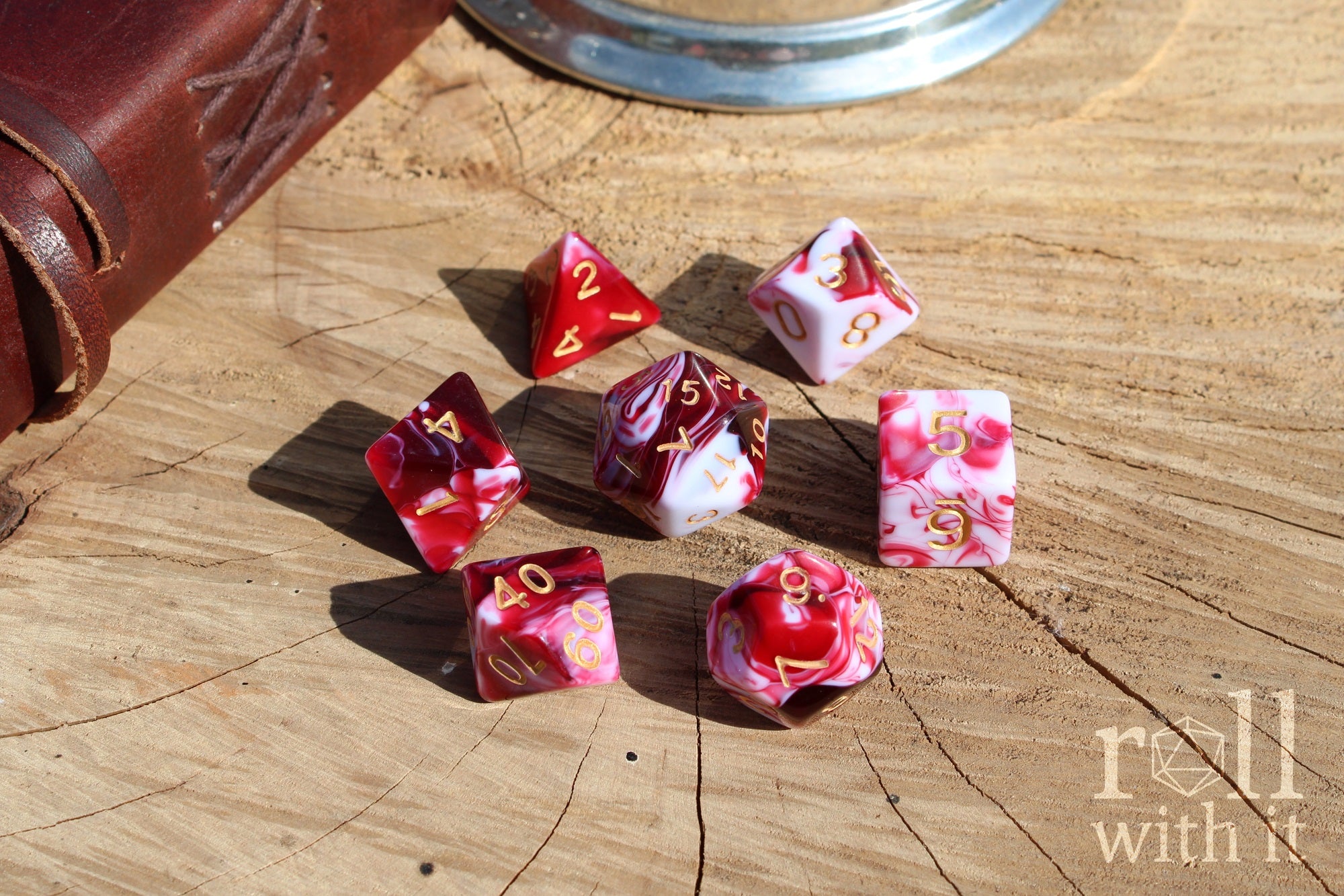 A set of 7 cherry red and swirling creamy white polyhedral TTRPG dice on a oak base with role playing game accessories in the background