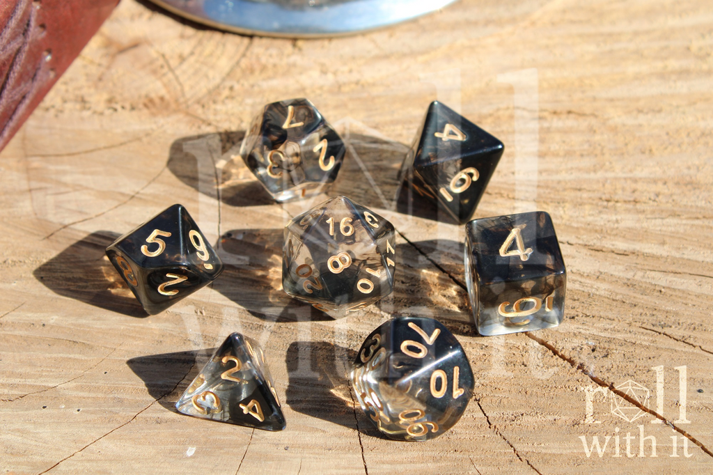 A set of smoky black and clear polyhedral roleplaying dice with gold numbering sitting on an oak surface