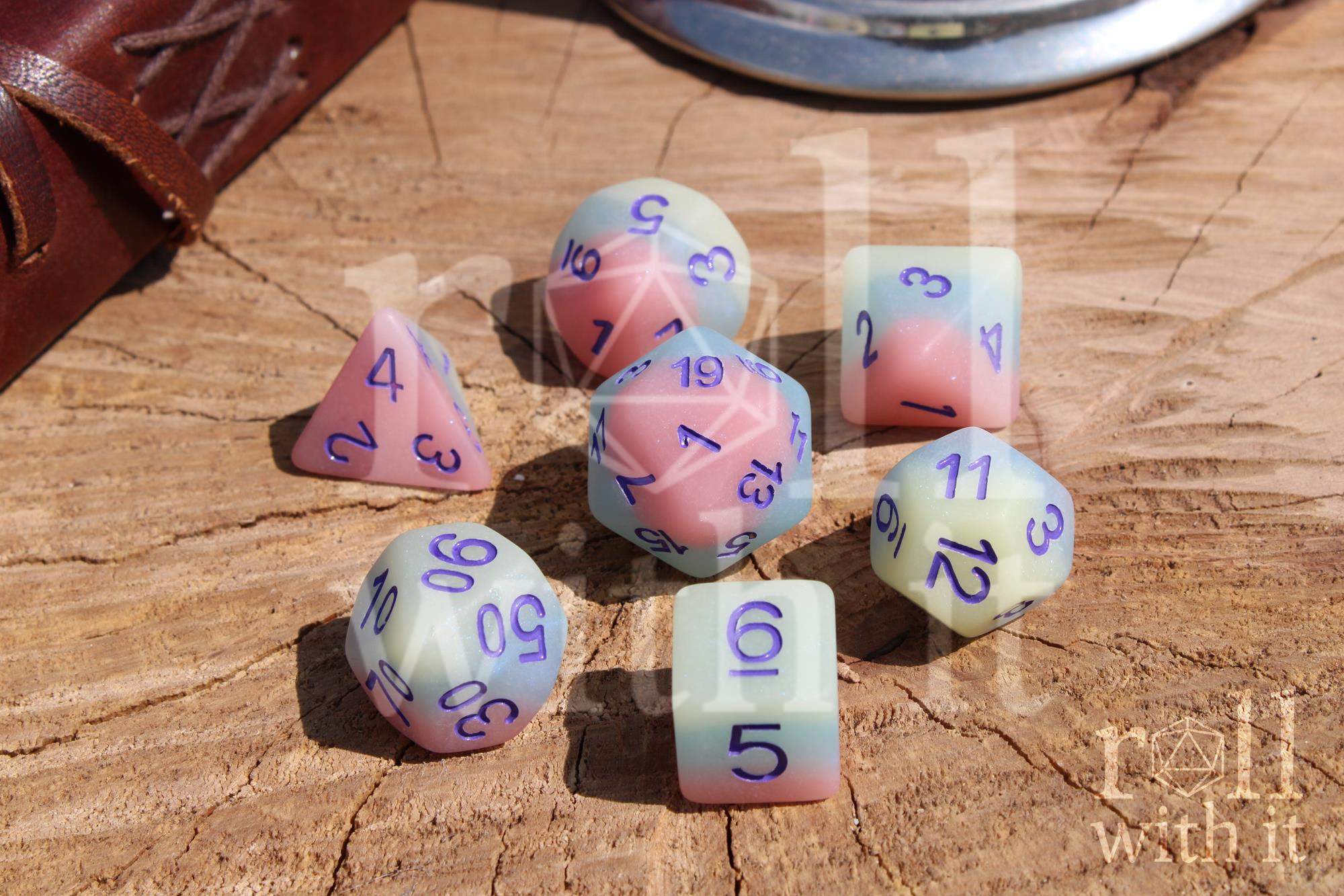 Set of matt pastel-coloured polyhedral dice with purple numbers on a wooden surface, featuring the text 'roll with it'.