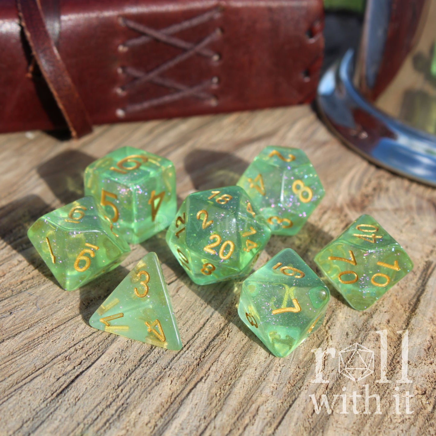 Green glittery clear polyhedral dice set with gold numbers on a wooden surface, with 'roll with it' text.