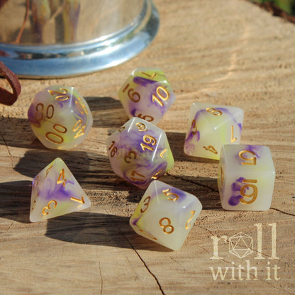 Set of 7 polyhedral dice with purple and gold numbers on a wooden surface, featuring the brand 'roll with it'.