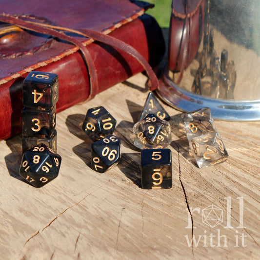 Set of 11 clear and smoky black polyhedral roleplaying dice on a wooden surface with tabletop RPG accessories in the background
