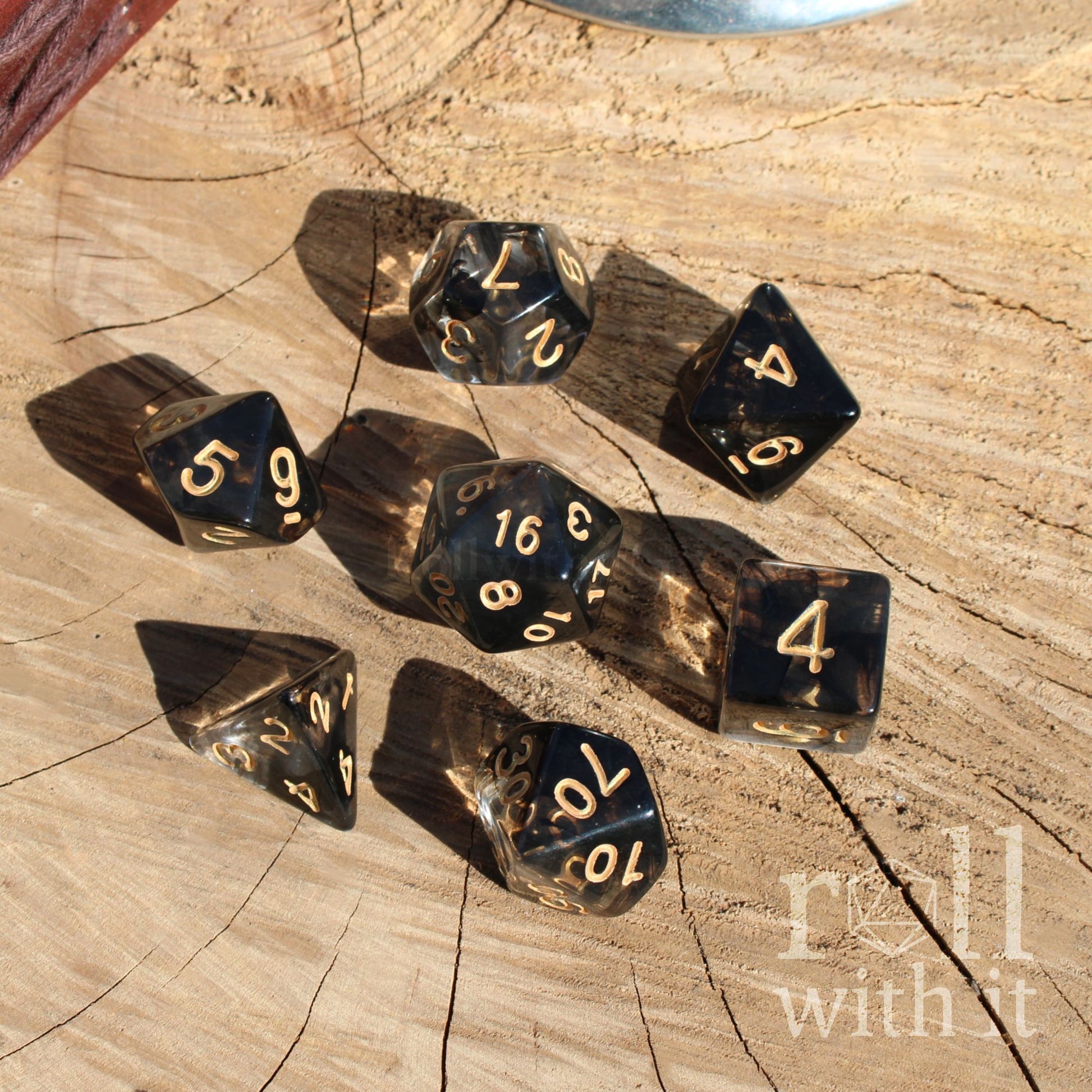 Set of clear and smoky black polyhedral roleplaying dice on a wooden surface with tabletop RPG accessories in the background