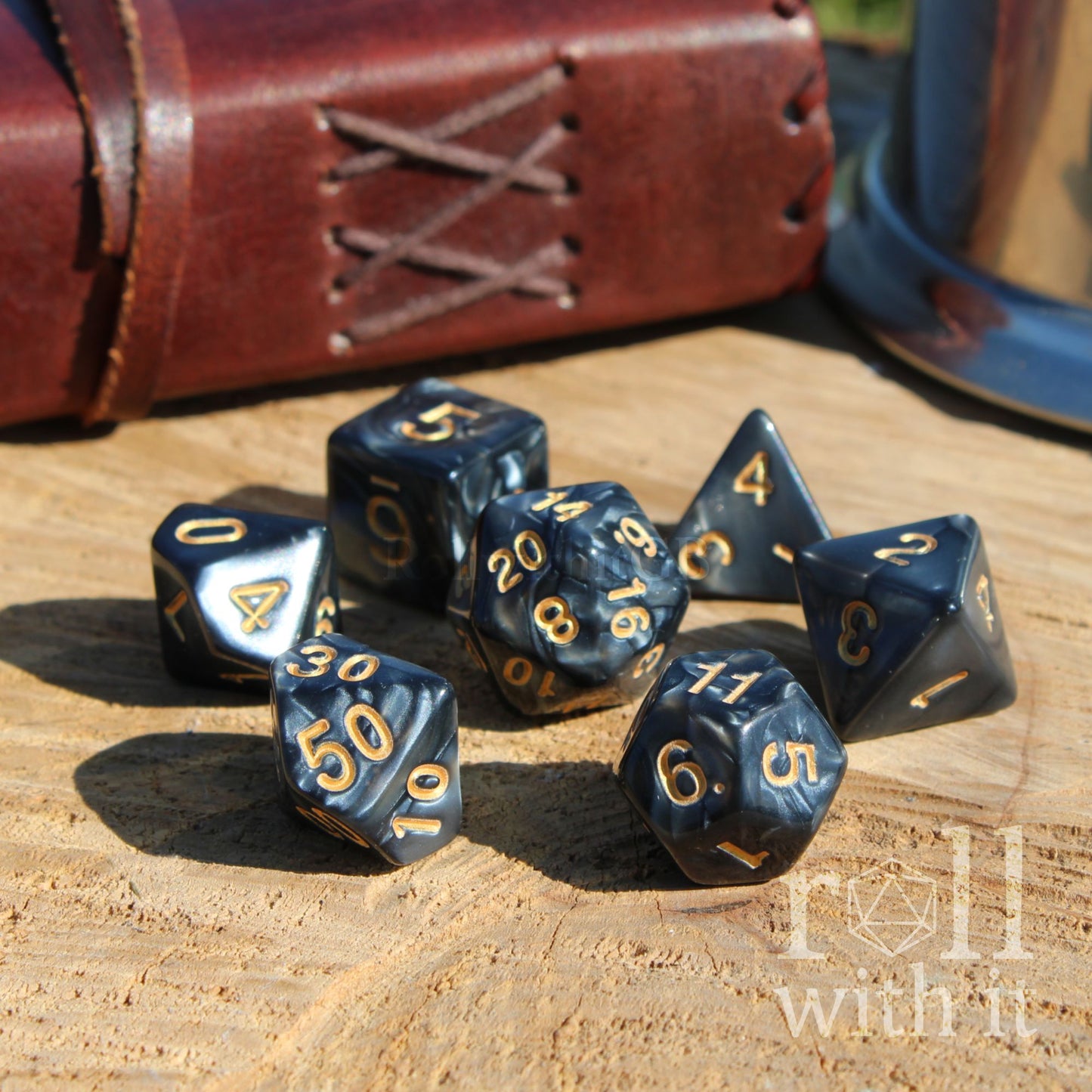 Set of black polyhedral dice with gold numbers on a wooden surface, with a leather-bound book in the background.