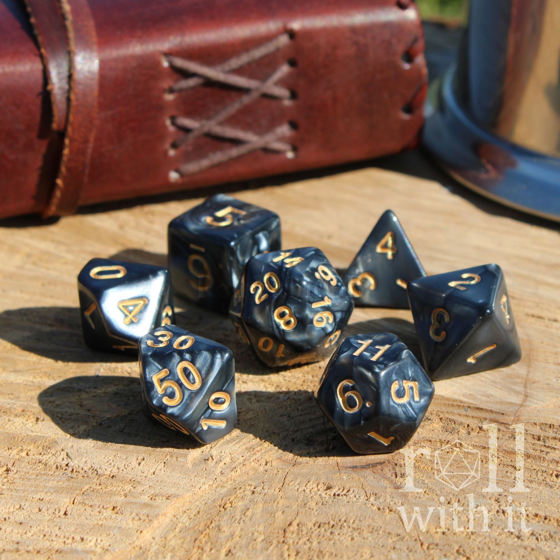 Set of black polyhedral dice with gold numbers on a wooden surface, with a leather-bound book in the background.