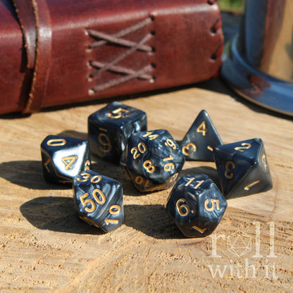Set of black polyhedral dice with gold numbers on a wooden surface, with a leather-bound book in the background.