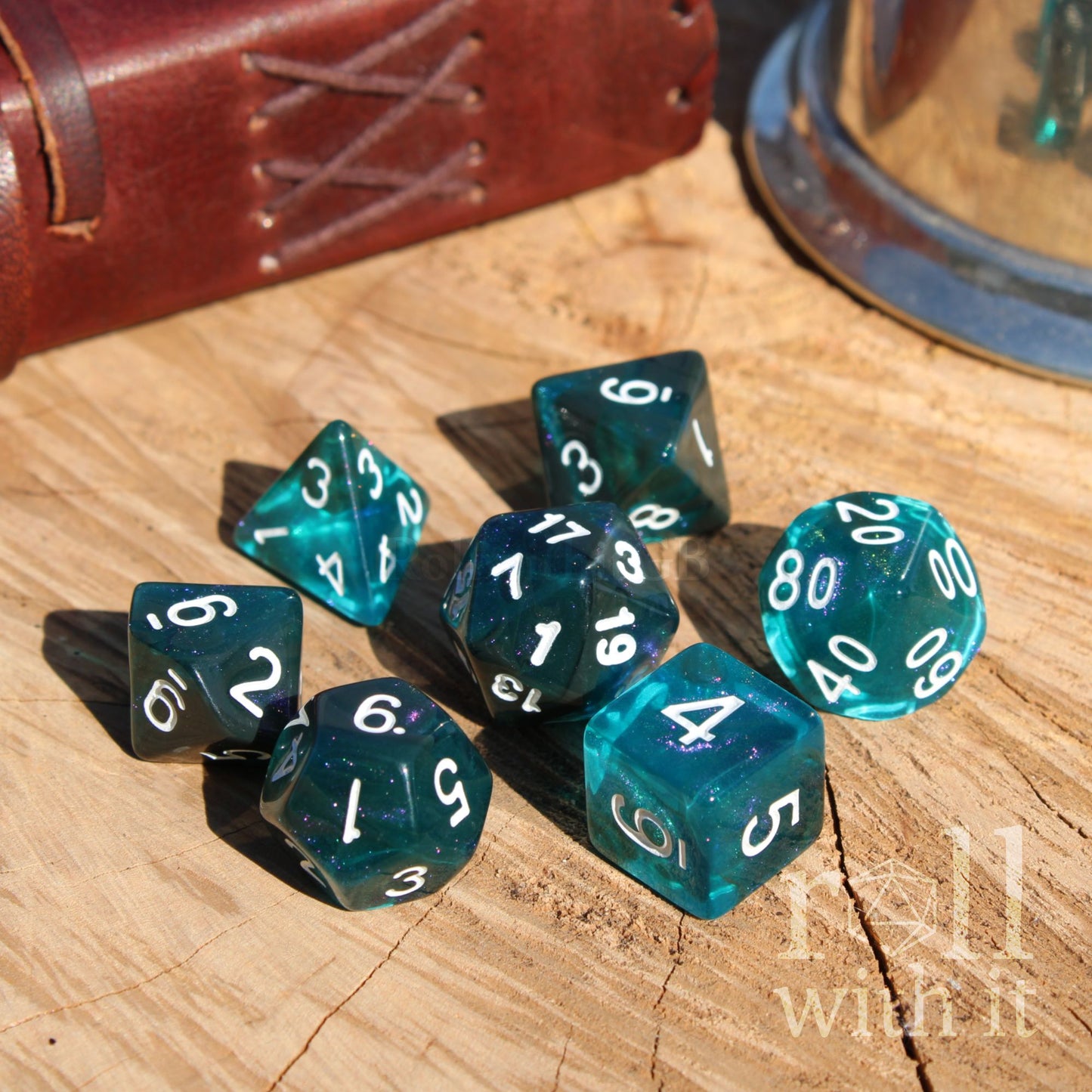Set of teal polyhedral dice with white numbers on a wooden surface, with a leather book in the background.