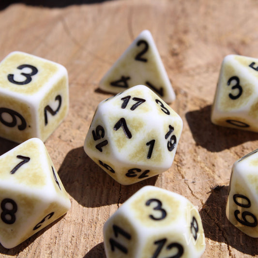 Set of 7 creamy bone white matte D&D roleplaying dice with a worn looking surface, with black numbers on a wooden surface