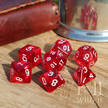 7 saturated colourful cherry red D&D dice with white numbers on a wooden surface, with a leather book in the background