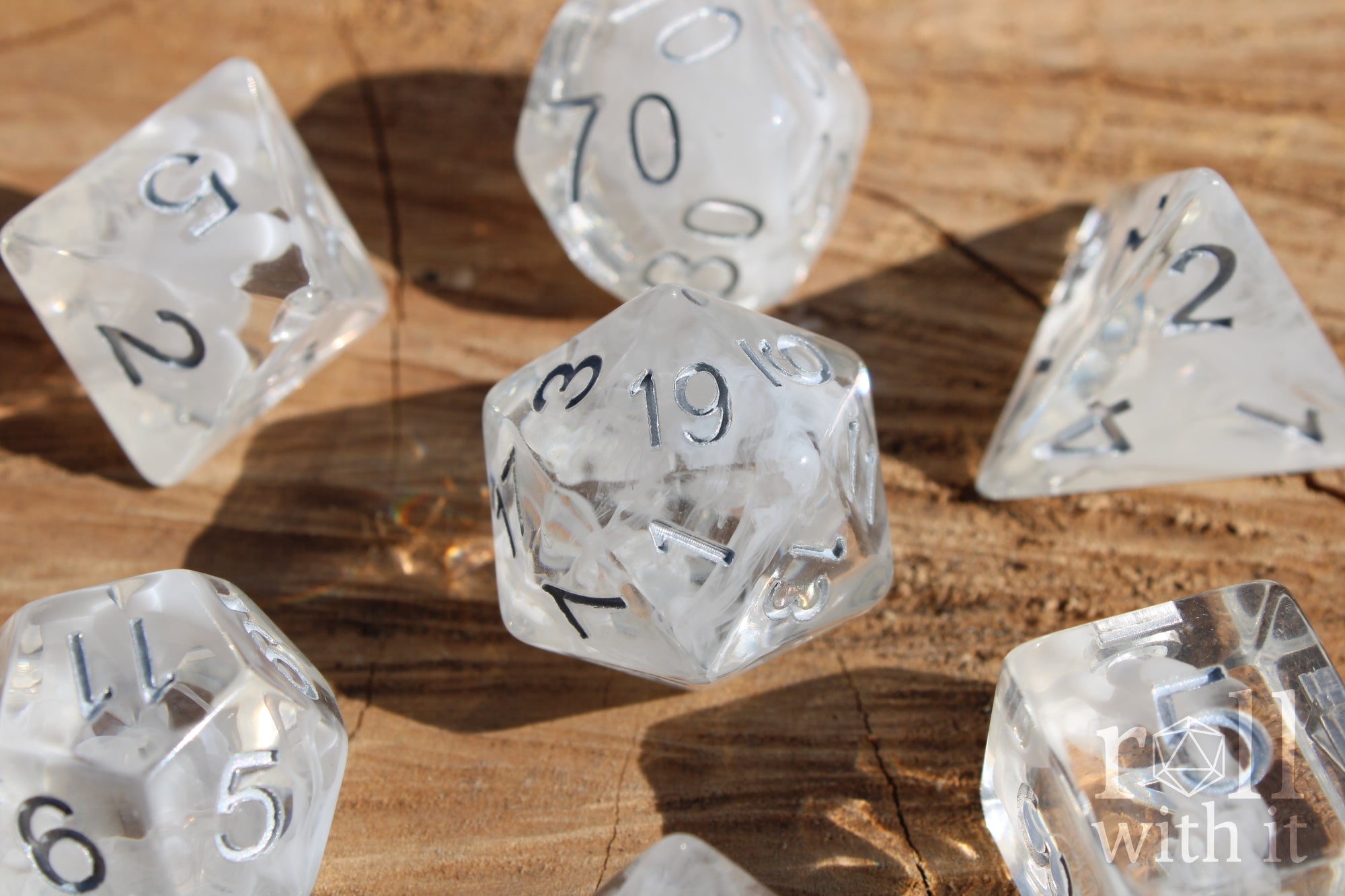 A set of 7 clear and white smoky polyhedral roleplaying dice with silver numbers on a wooden surface