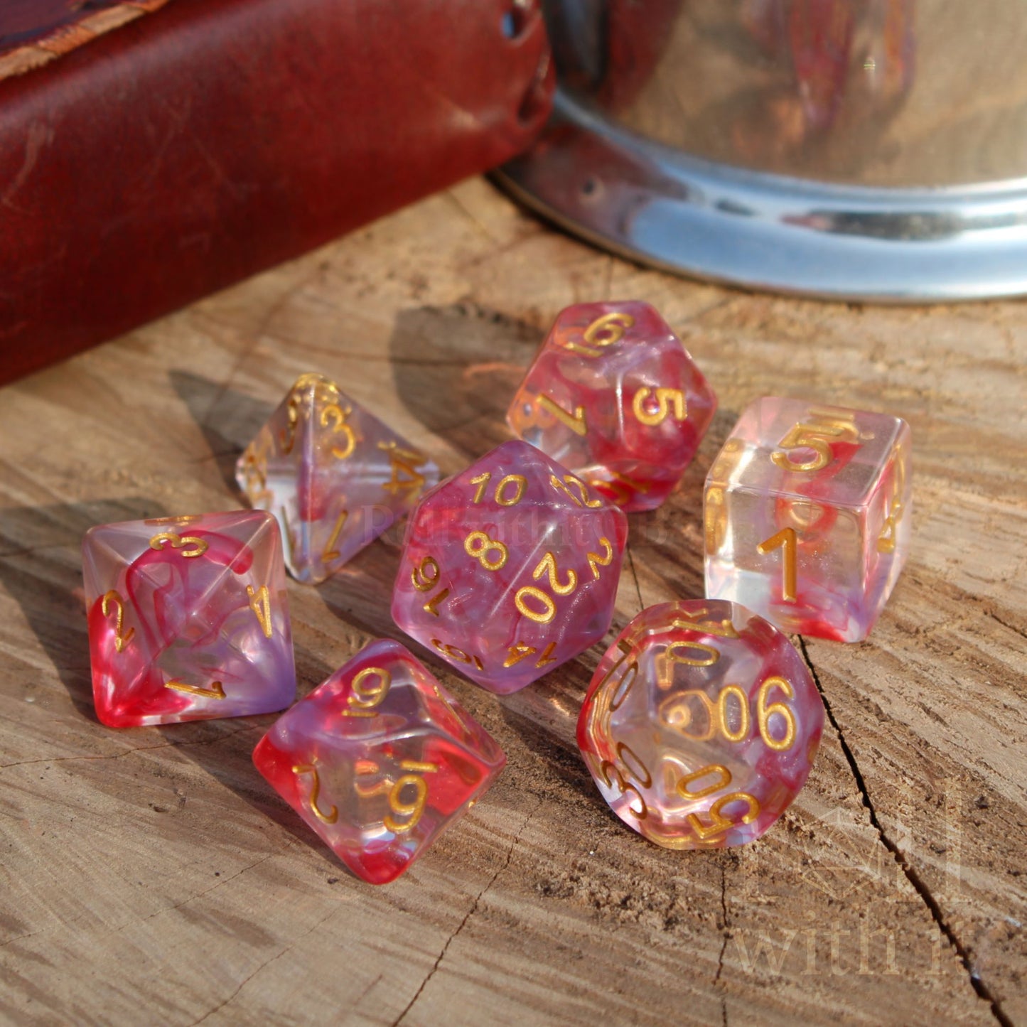 Set of 7 rose pink and clear polyhedral D&D dice with gold numbers on a wooden surface.