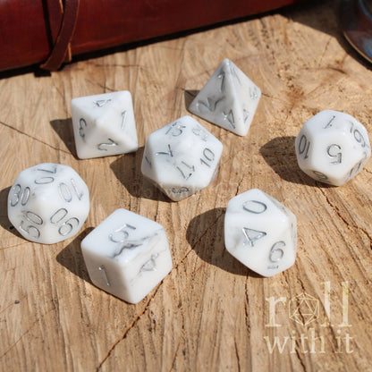 A side view of 7 resin white marble effect luxury polyhedral tabletop roleplaying dice on a wooden surface with silver numbers