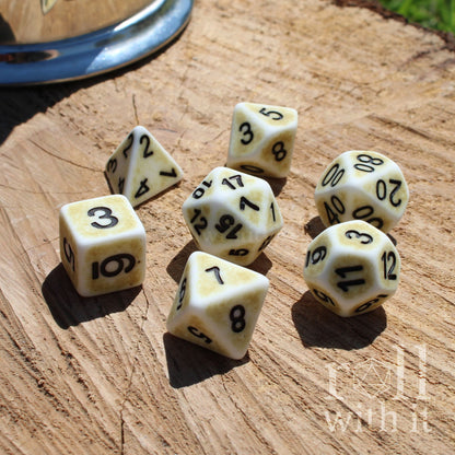 Angled view of 7 polyhedral white bone roleplaying TTRPG dice on a wooden surface with black easy to read letters