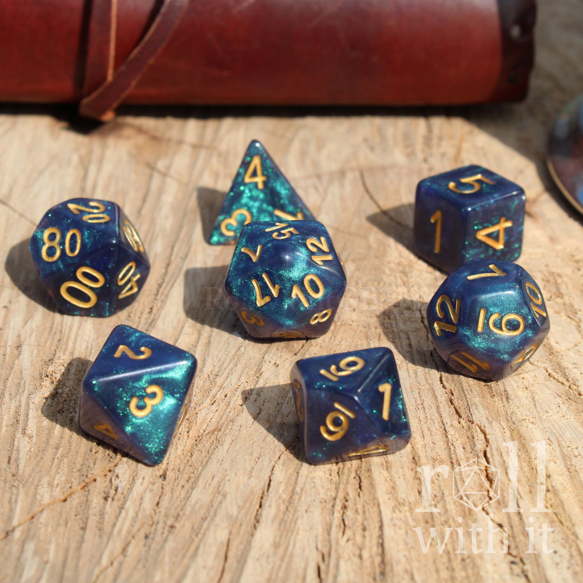 Set of 7 polyhedral blue glitter D&D polyhedral dice with gold numbers on a wooden surface, with 'roll with it' text.
