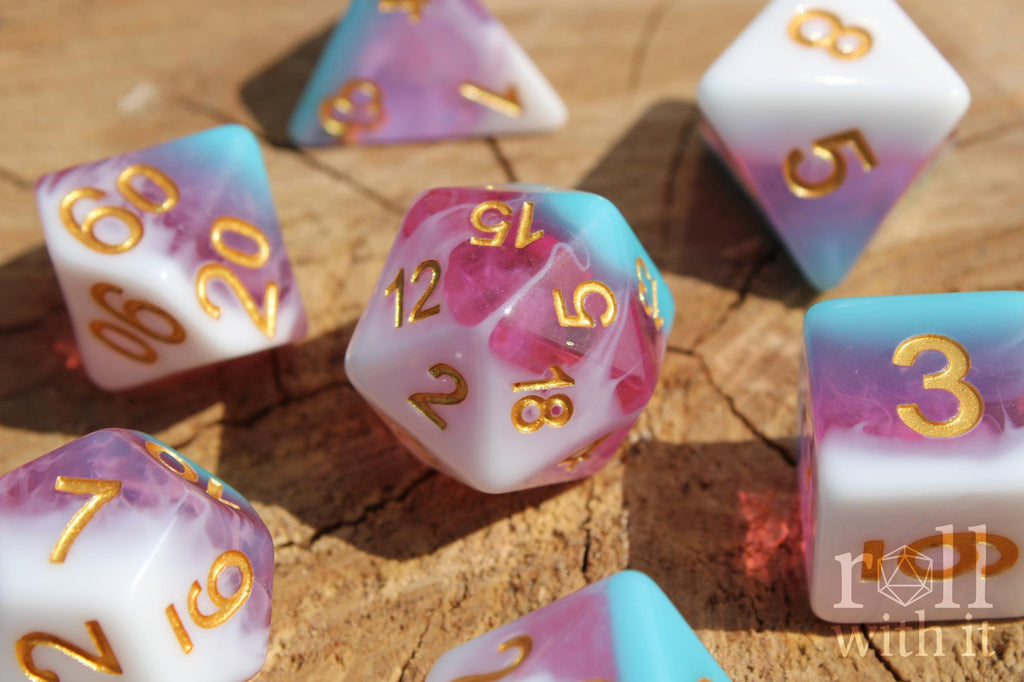 Set of colourful layered pink, light blue, and creamy white polyhedral roleplaying dice with gold numbers on a wooden surface