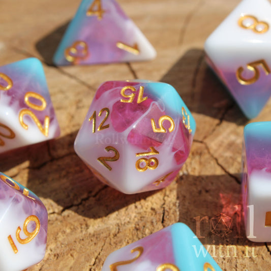 Set of colourful layered pink, light blue, and creamy white polyhedral roleplaying dice with gold numbers on a wooden surface