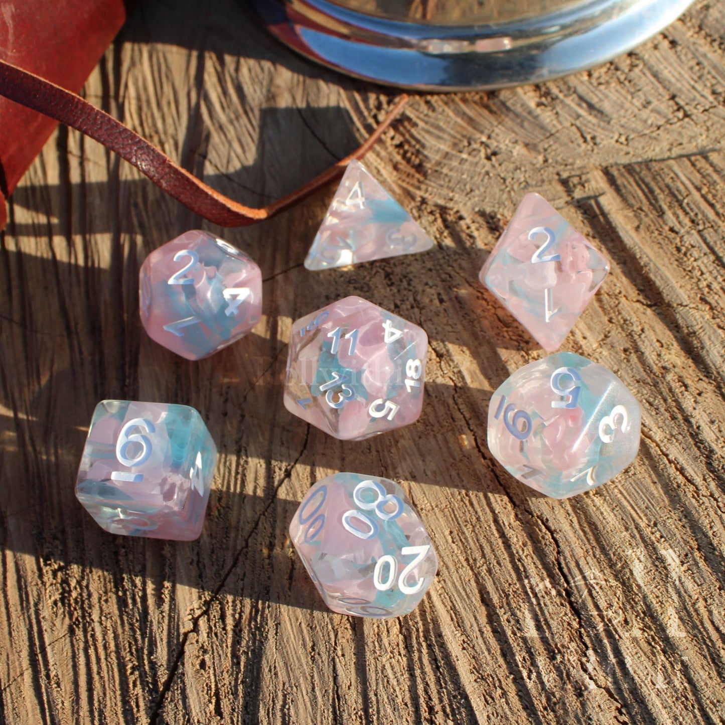 Set of 7 cute pale pink and blue ribbon swirling fantasy aesthetic D&D roleplaying game dice set on a wooden surface with white numbers