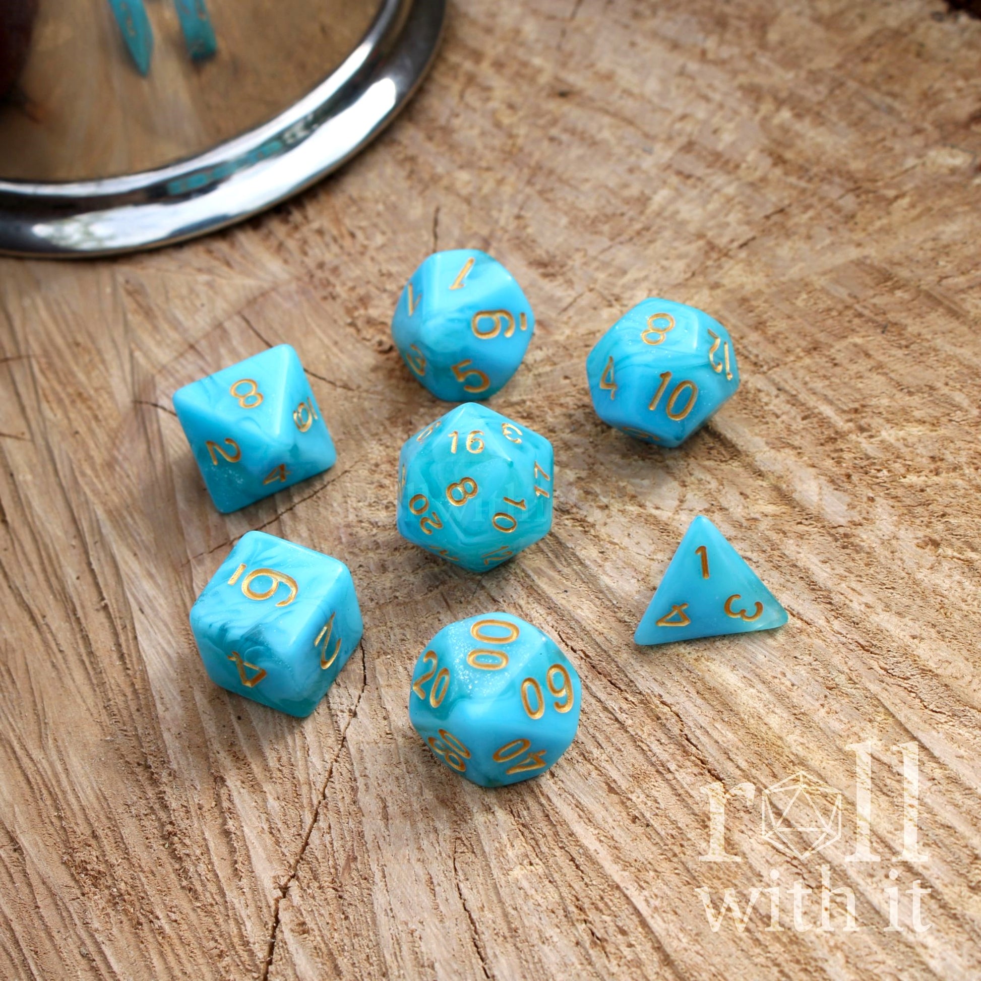 Set of 7 bright fresh blue polyhedral dice with gold numbers on a wooden surface, featuring the brand 'roll with it'.