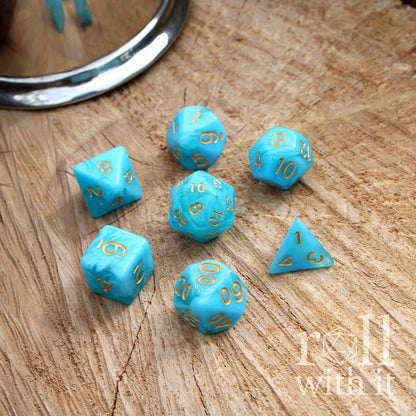 Set of 7 bright fresh blue polyhedral dice with gold numbers on a wooden surface, featuring the brand 'roll with it'.