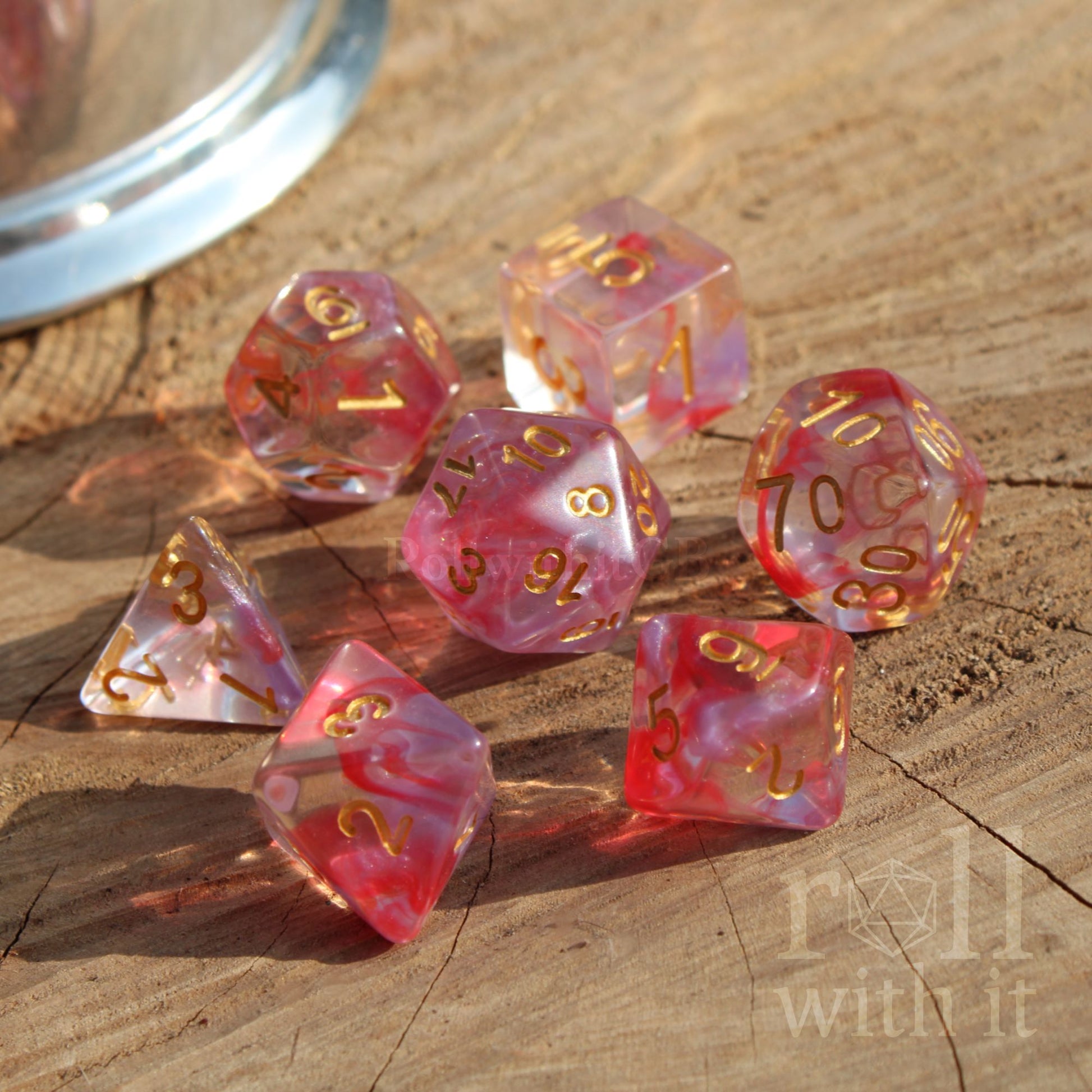 Set of 7 cute pastel pink and clear dice with gold numbers on a wooden surface, featuring the text 'roll with it'.
