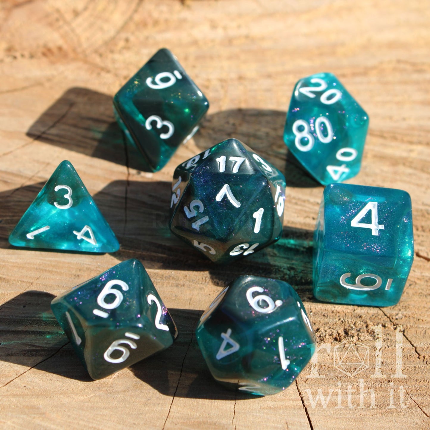 Set of dark teal polyhedral dice with white numbers on a wooden surface, featuring the brand 'Roll with It'.