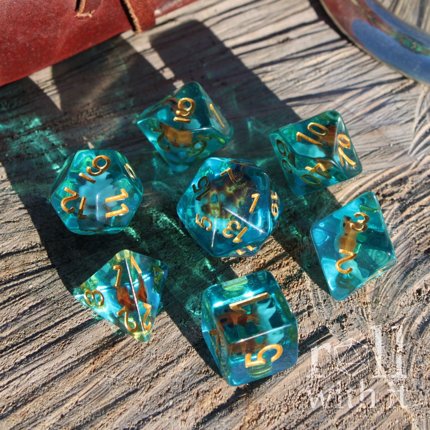 Set of clear blue turquoise polyhedral dice with gold numbers and a mini goldfish inside on a wooden surface