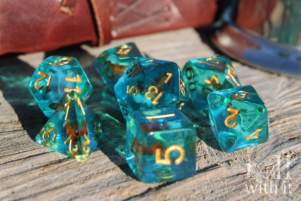 Set of clear blue turquoise polyhedral dice with gold numbers and a mini goldfish inside on a wooden surface