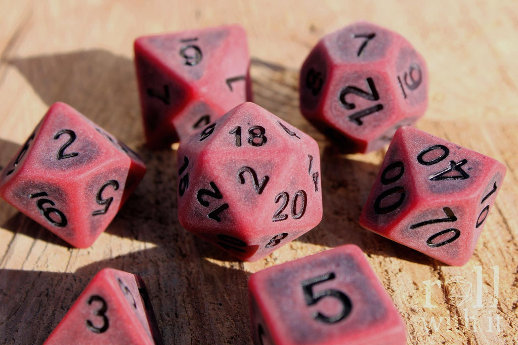 Matte red polyhedral roleplaying dice with a worn look with black lettering set on a wooden surface