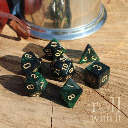 Set of 7 emerald glitter filled nebula green polyhedral dice with gold numbers on a wooden surface, with 'roll with it' text.