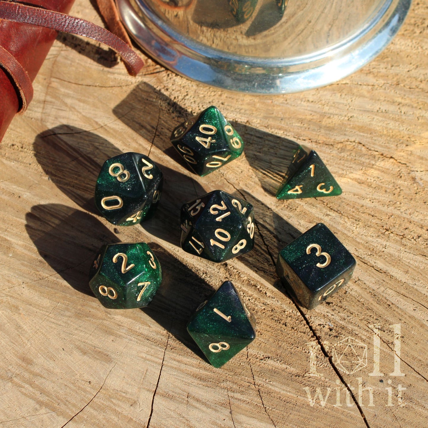 Set of 7 emerald green galaxy glitter polyhedral dice set on a wooden surface with a metallic cup in the background.