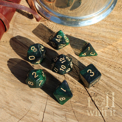 Set of 7 emerald green galaxy glitter polyhedral dice set on a wooden surface with a metallic cup in the background.