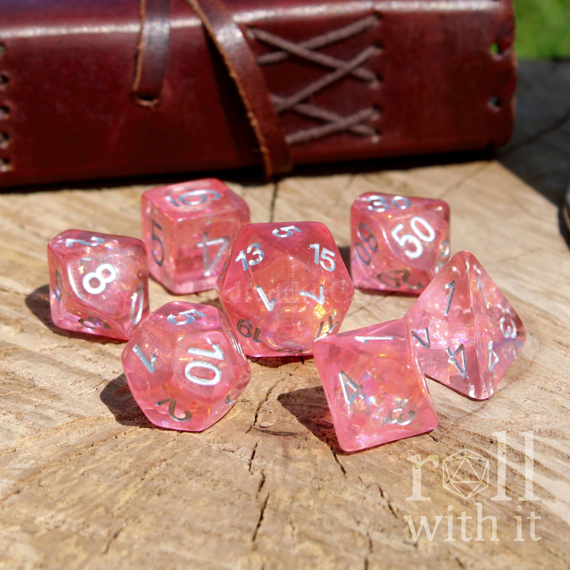 Set of foil pink dice with white numbers on a wooden surface, with a leather-bound book in the background.