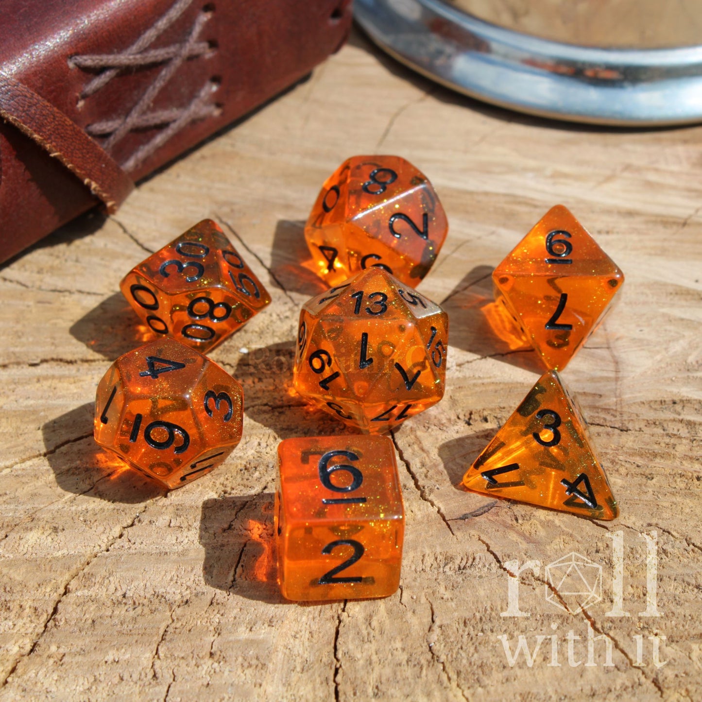 Set of bright glitter filled orange dice with black numbers on a wooden surface, with a leather-bound book and metal object in the background.