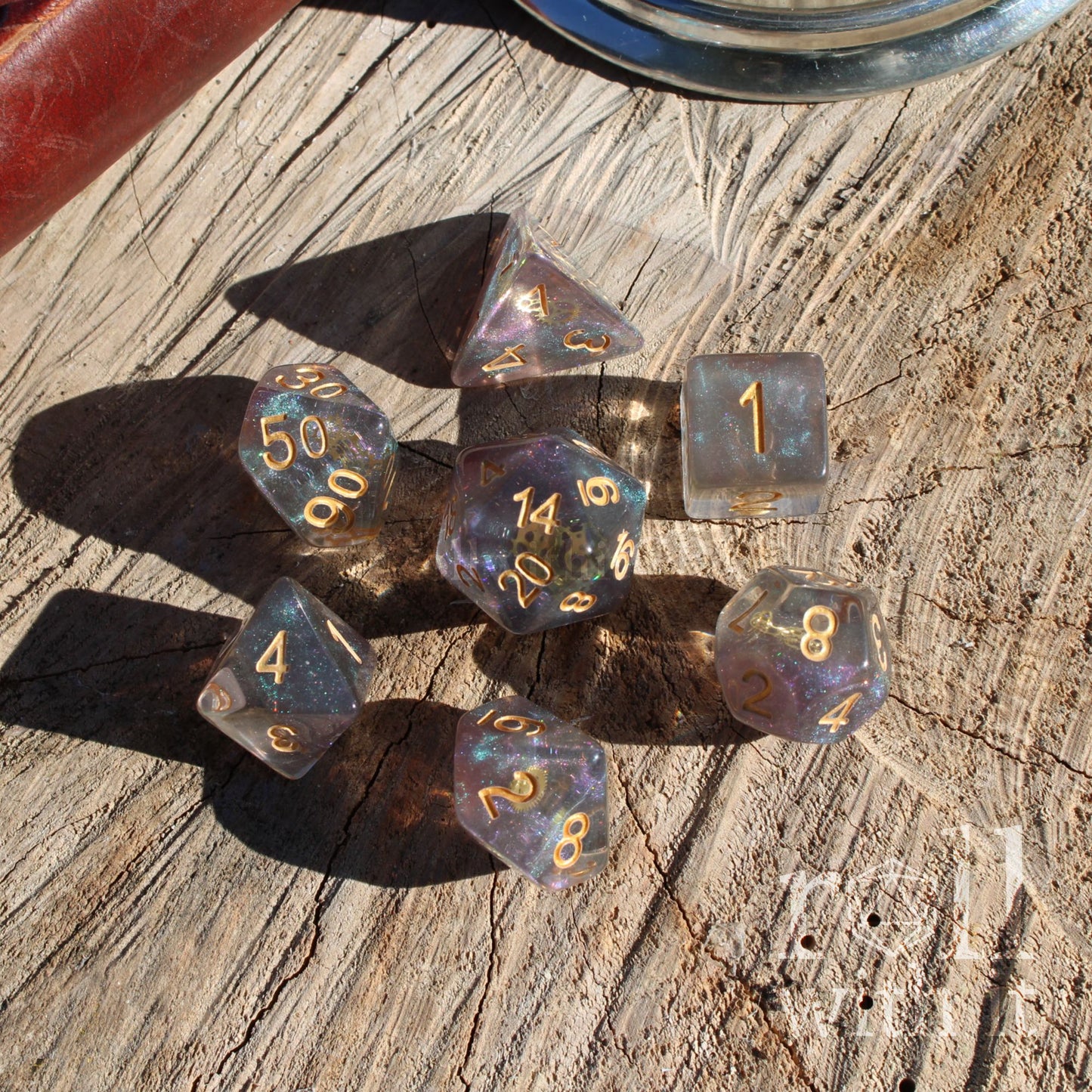 Set of 7 polyhedral roleplaying dice made of glitter filled resin with golden inked numbers and with golden gears inside on a wooden surface