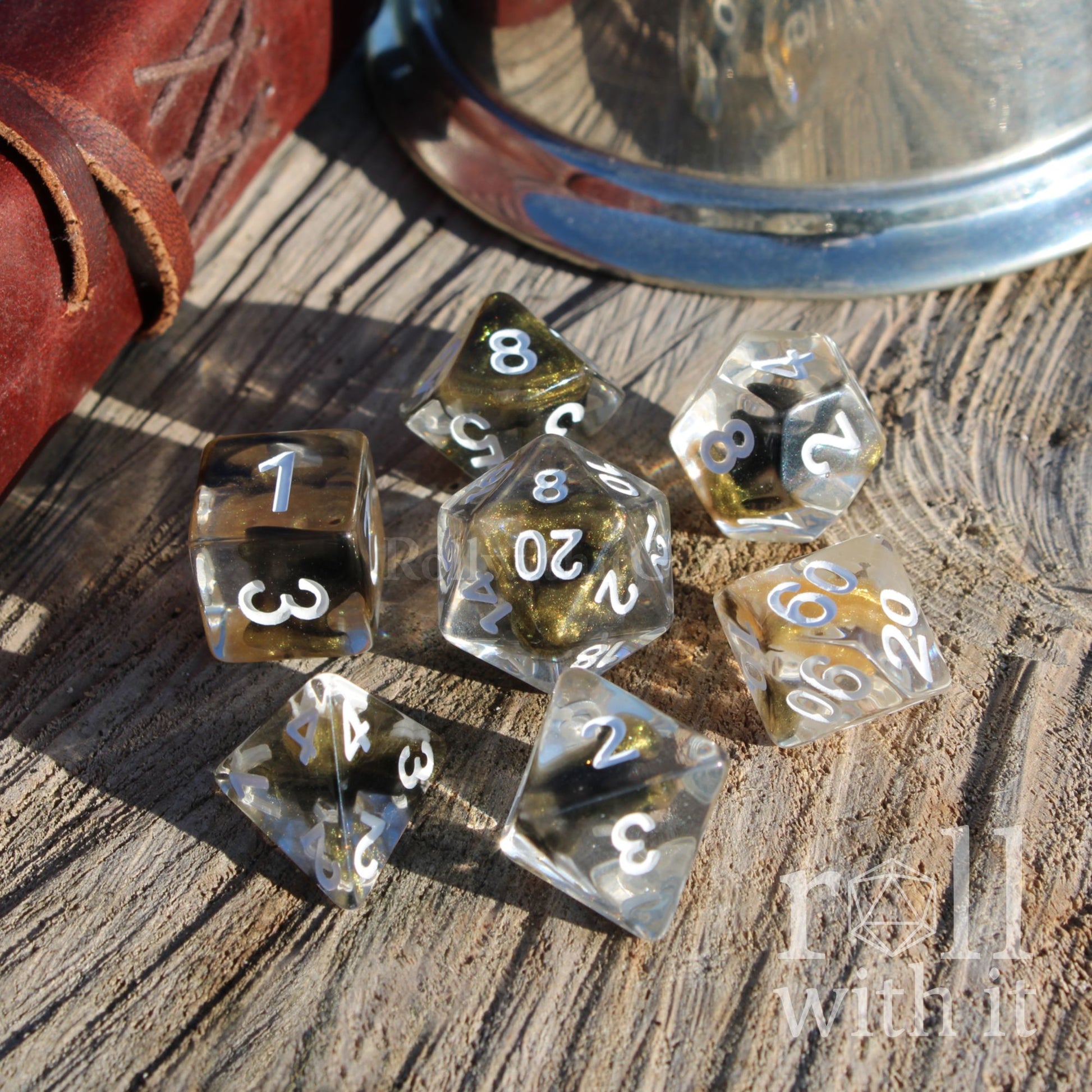 Set of glittering gold polyhedral dice on a wooden surface with a leather case and metal container in the background.