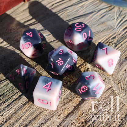 Angled view of 7 pink black polyhedral D&D dice on a wooden surface with pink numbers
